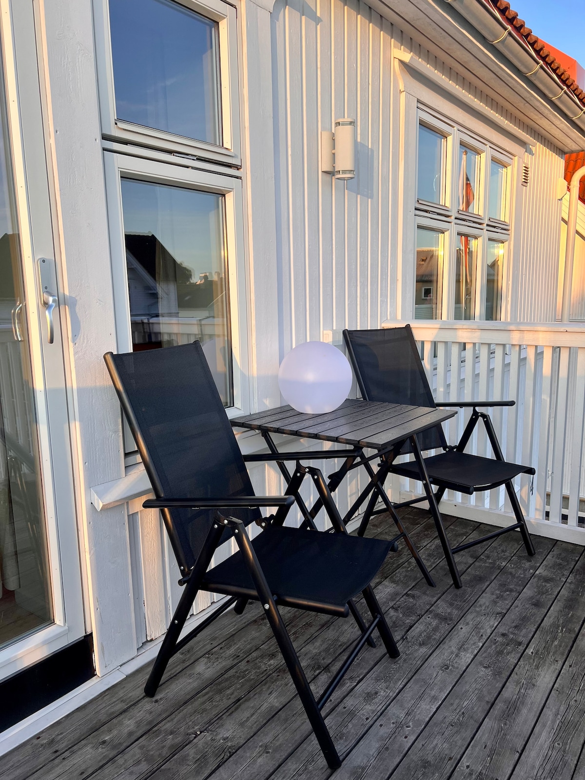 A private terrace features a small table with two folding chairs, providing an inviting space to relax. A round light fixture adds a soft glow, while sunlight reflects off the nearby windows, enhancing the serene atmosphere of the setting.