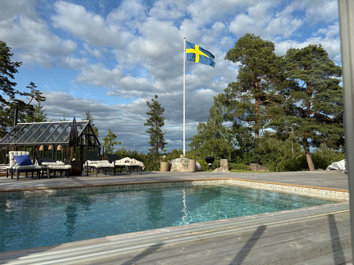 A serene outdoor pool invites relaxation, framed by a wooden deck. Nearby, a Swedish flag stands prominently against a sky filled with clouds. Comfortable lounge chairs are positioned around the pool, with a glasshouse visible in the background amid surrounding trees.