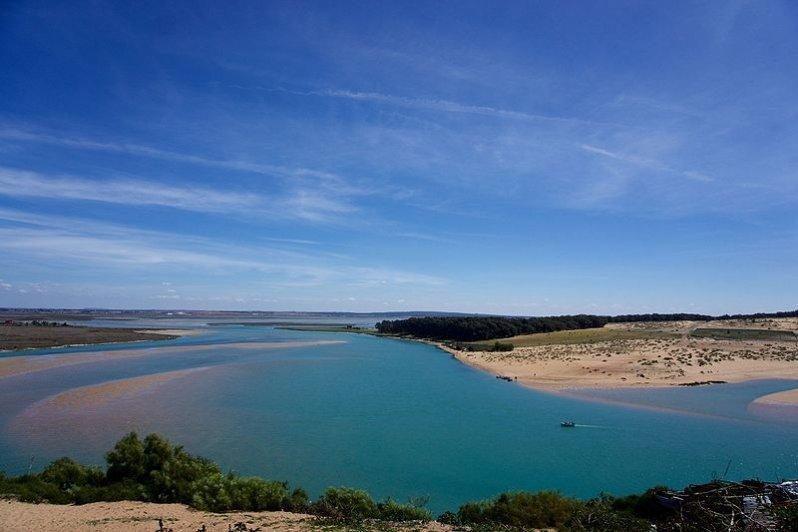 A panoramic view of a river winding through a landscape of sandy shores and lush greenery. The clear blue water reflects the sky, while gentle waves create a textured surface. The horizon showcases a blend of blues and greens, suggesting a peaceful natural setting.