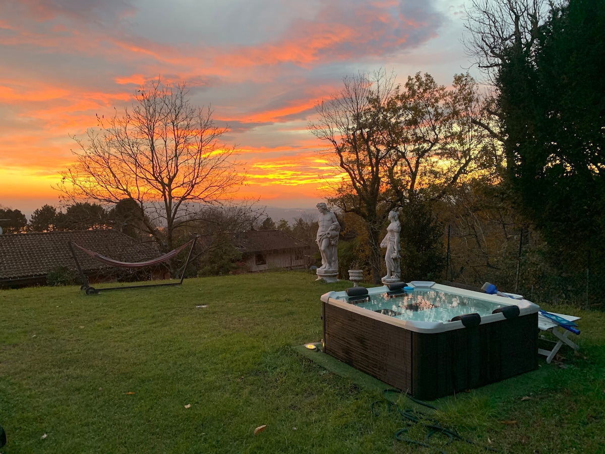 A serene outdoor space features a heated hot tub surrounded by a well-maintained lawn. Two elegant statues stand nearby, complemented by a hammock gently swaying in the foreground. The sky displays a warm gradient of orange and pink hues as the sun sets.