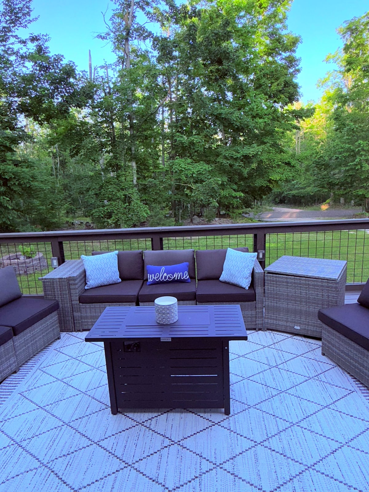 A cozy outdoor seating area is arranged with dark wicker furniture and soft cushions. A central coffee table is positioned on a geometric-patterned rug. A decorative pillow with the word 'welcome' rests on one of the chairs. Lush trees provide a natural backdrop.