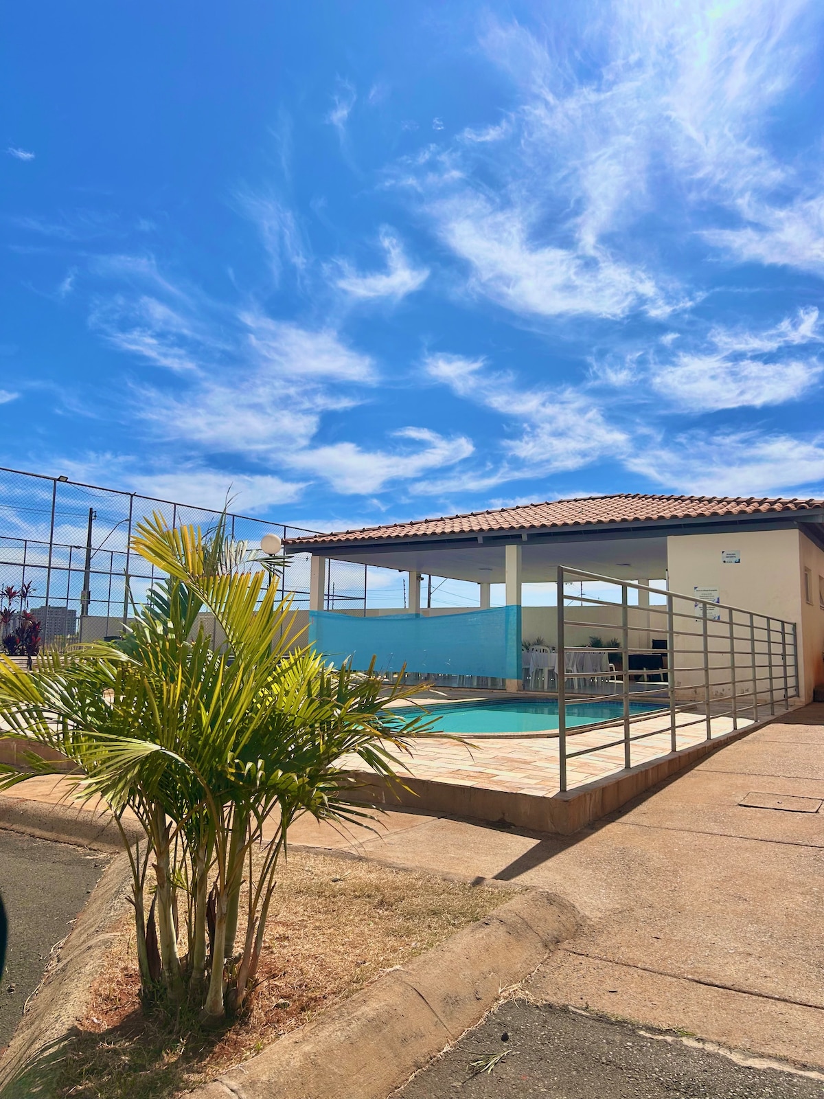 A well-maintained outdoor space features a swimming pool surrounded by a secure railing. A covered area can be seen nearby, with a clear sky above and a palm plant positioned in the foreground, adding a touch of greenery to the scene.