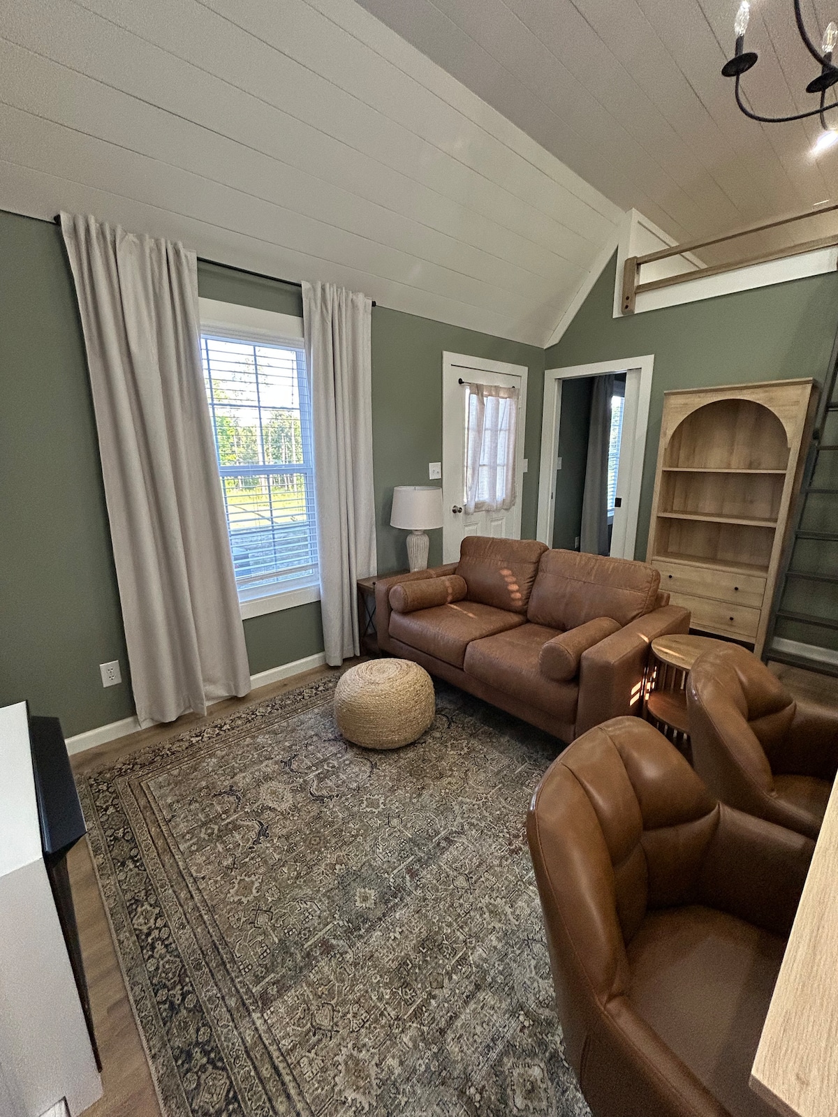 A cozy living area features a brown leather sofa and two armchairs, complemented by a natural fiber pouf. Soft, neutral curtains frame a window allowing natural light, while a small table lamp adds illumination. A built-in bookcase and staircase are visible in the background.
