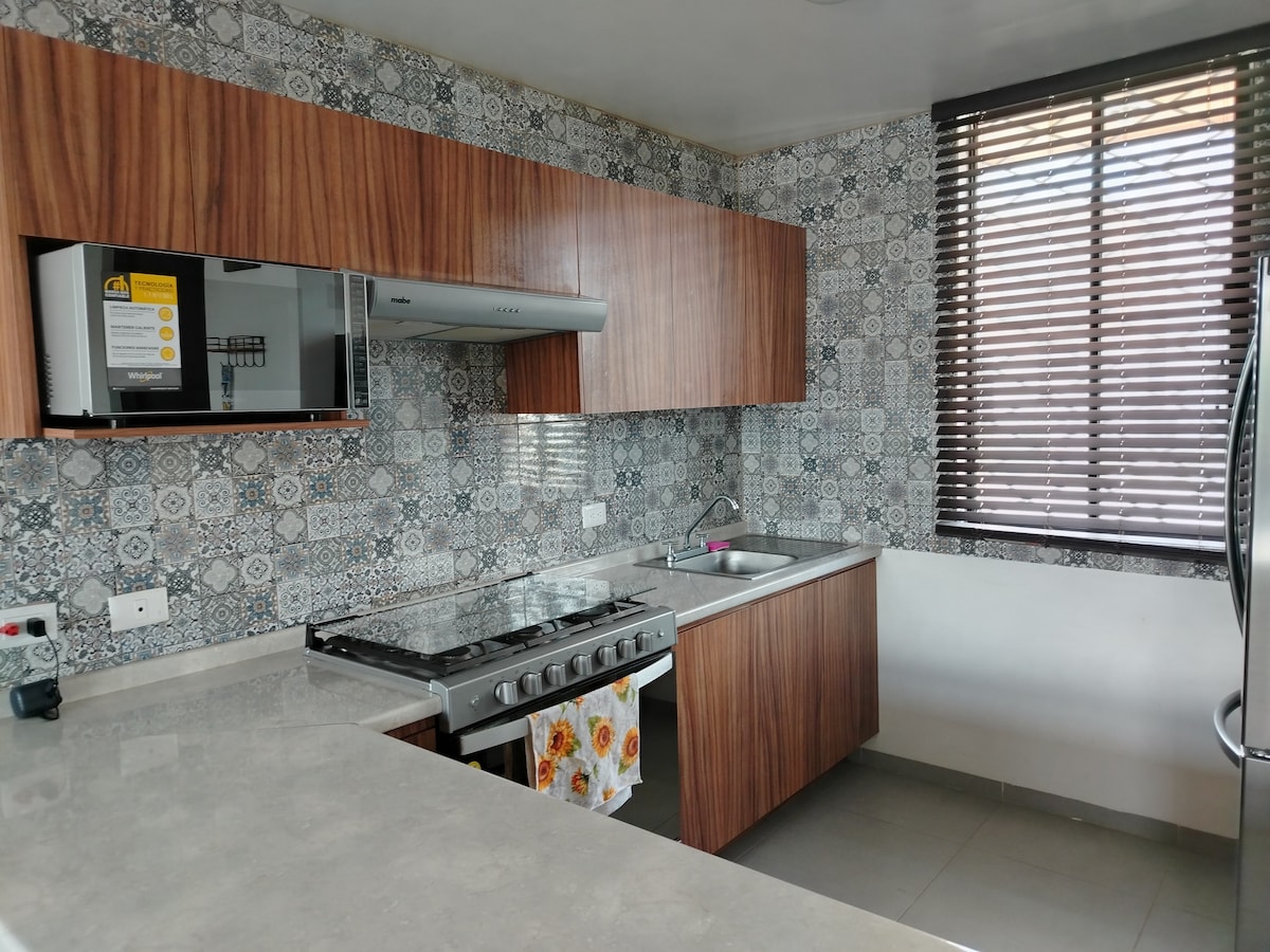 A modern kitchen features wooden cabinetry and a tile backsplash with intricate patterns. Stainless steel appliances include a gas stove and a microwave. Natural light filters through horizontal blinds, illuminating the workspace, which is completed by a stone countertop and a sink.