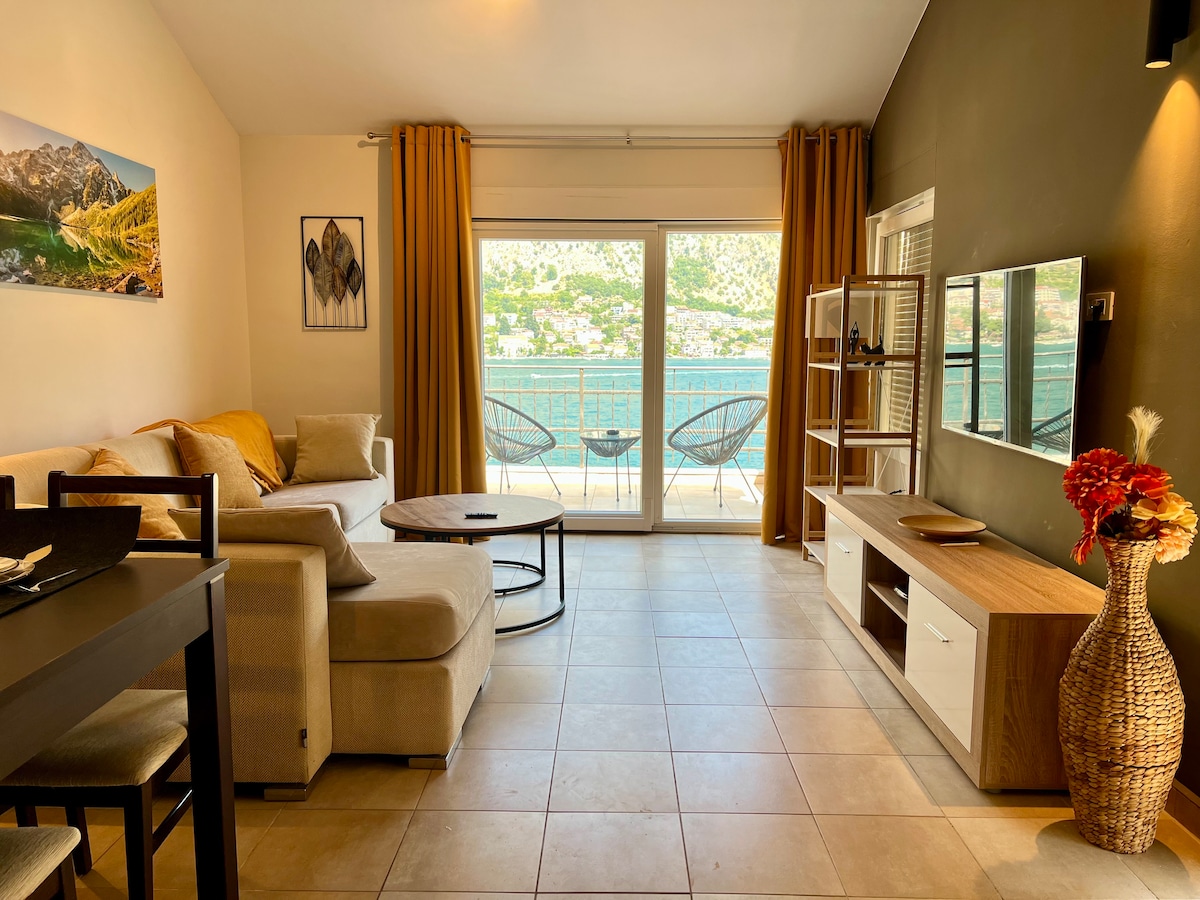 A stylish living area features a comfortable seating arrangement with a light-colored sofa and accent pillows. Large glass doors open to sea views, complemented by warm curtains. A round coffee table and a modern entertainment unit provide functionality. Decorative elements add a touch of character.