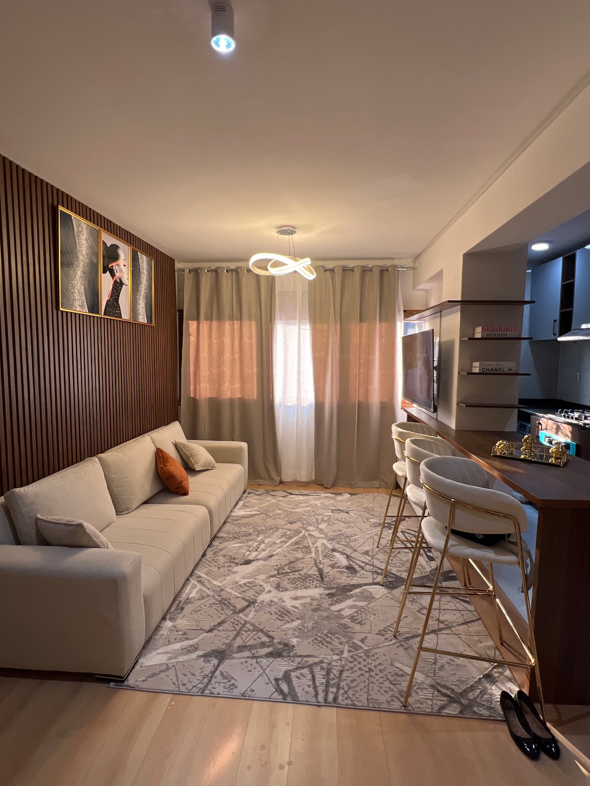 A living area features a light-colored sofa with a decorative pillow, paired with stylish bar stools at a counter. Natural light flows through sheer curtains, which frame a modern light fixture and showcase wooden paneling on one wall. A patterned rug adds texture to the space.