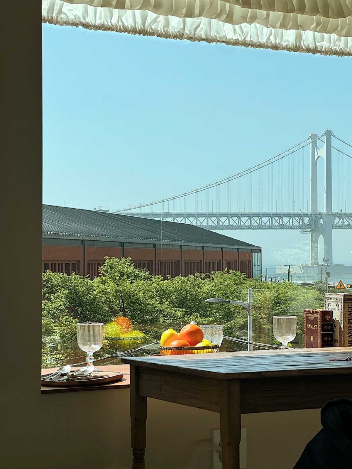 A scenic view from the window showcases a bridge spanning over calm waters, framed by lush greenery. A table is positioned nearby, adorned with glasses and a bowl of fruit, inviting a moment of relaxation while enjoying the vibrant surroundings.
