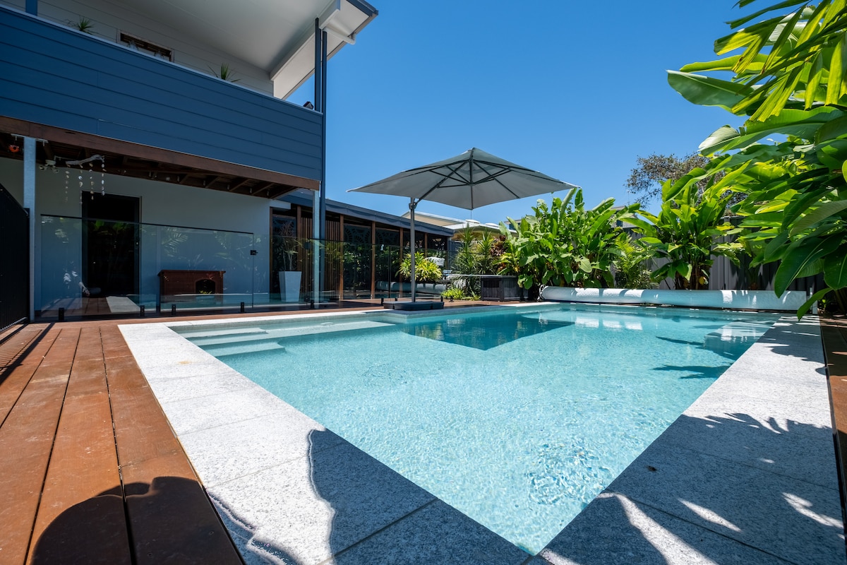 A refreshing swimming pool is surrounded by a wooden deck and lush greenery. A large umbrella provides shade over a seating area. The clear blue water reflects the sunny sky, creating a calm and inviting outdoor space.