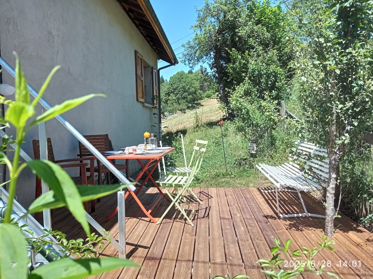 A wooden terrace is featured, furnished with a red table and several chairs in varying colors. A small table holds drinks and snacks, and surrounding greenery adds a touch of nature. The space invites relaxation with a view of the nearby landscape.