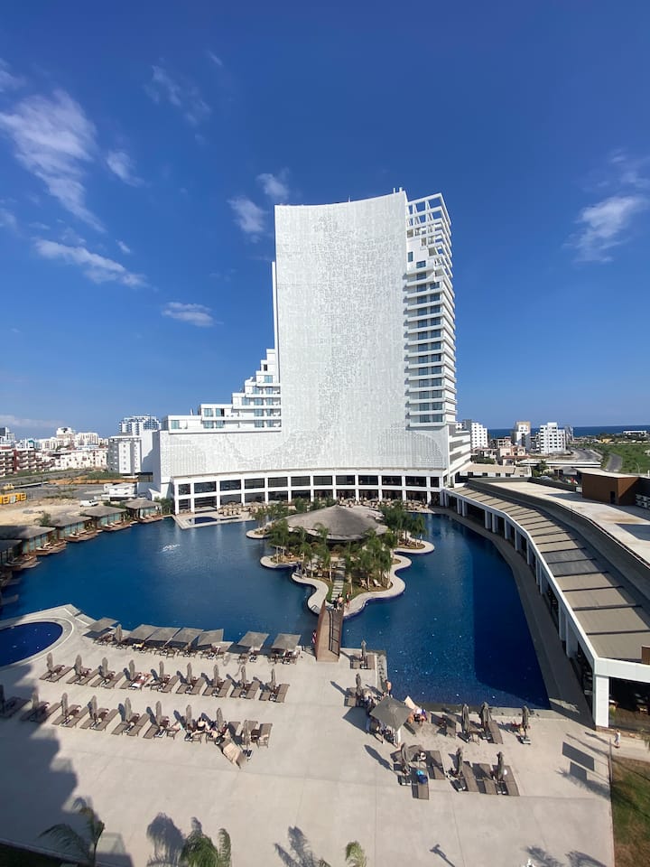 A large hotel building with a modern facade overlooks a spacious outdoor pool area. Sun loungers are arranged beside the pool, which is complemented by palm trees and a seating area. The blue sky enhances the overall inviting atmosphere of the resort.