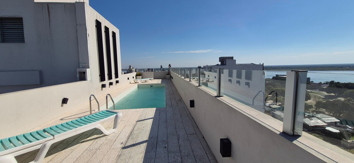 A rooftop pool area features modern lounge chairs alongside a clear swimming pool. Panoramic views of the skyline and landscape are visible beyond a glass railing, with bright blue skies overhead. Sunlight reflects off the water, creating a serene and inviting space.