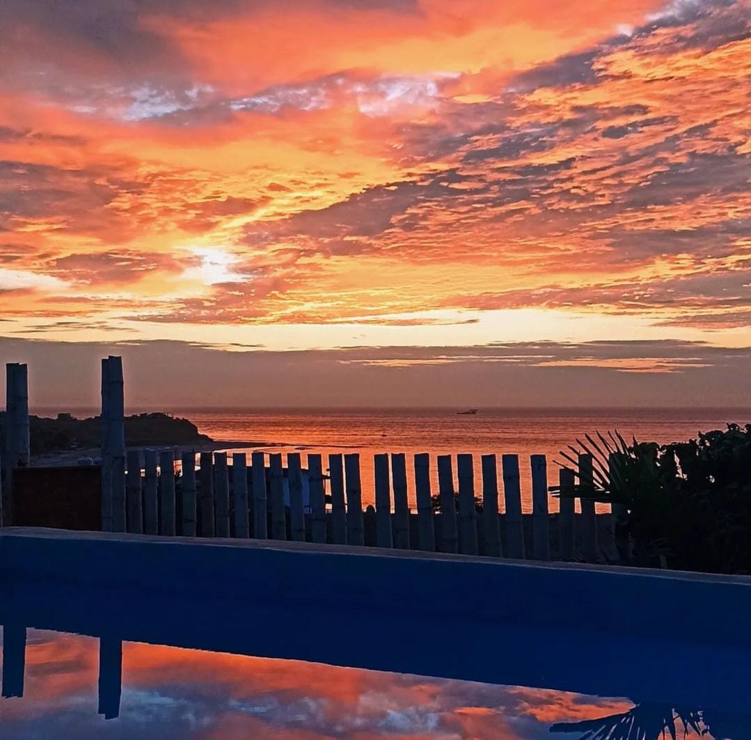 A serene sunset is captured over the ocean, with vibrant hues of orange and pink reflected in a pool. A wooden fence and tropical plants frame the scene, creating a peaceful atmosphere where the sky meets the water.