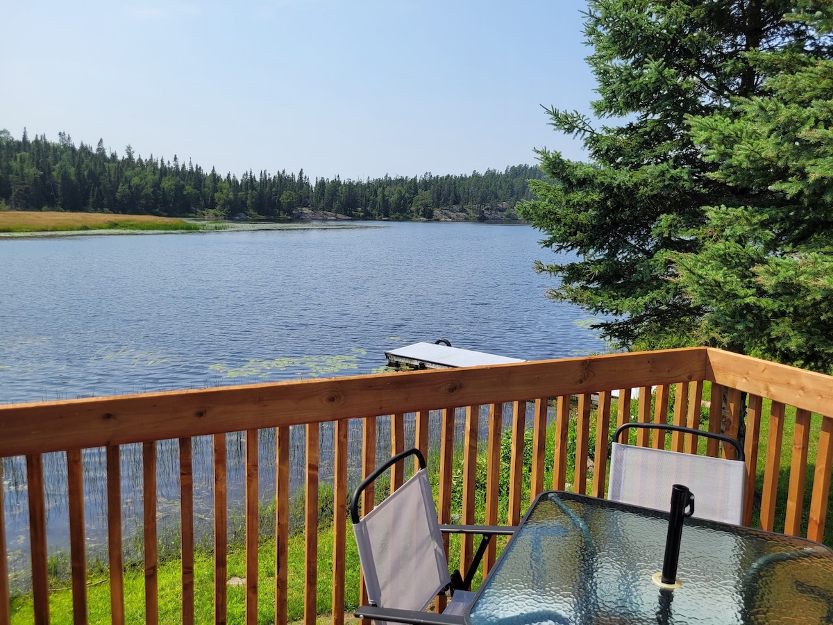 A wooden deck offers a clear view of the calm lake, framed by trees and distant hills. Two white chairs are positioned around a glass table, inviting relaxation. The water reflects the blue sky, creating a serene setting for outdoor enjoyment.