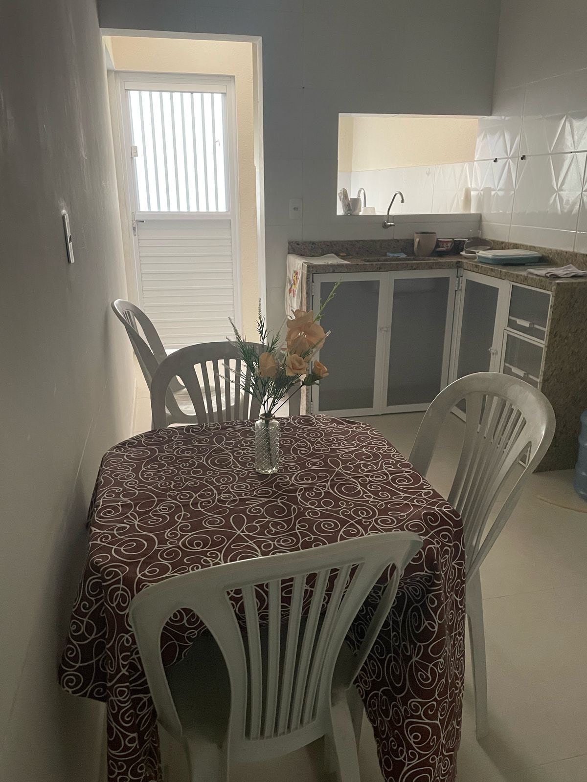 A small dining area features a round table covered with a patterned tablecloth and four white chairs. A vase with flowers is placed at the center of the table. A doorway leads to a kitchenette, where light streams in from a nearby window.