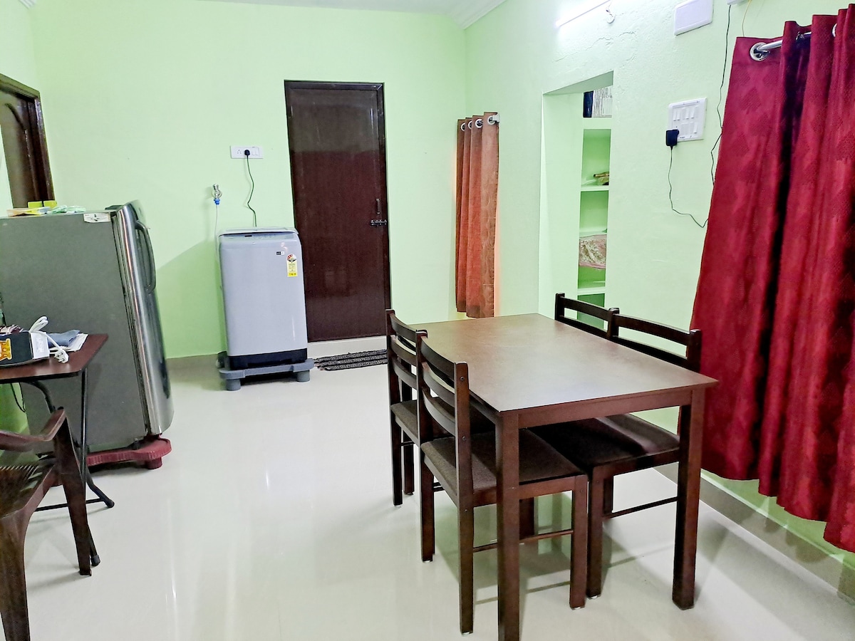 A living area features a simple wooden dining table surrounded by four chairs. In the background, a washing machine and a refrigerator are positioned near a door. Soft red curtains drape over a window, complementing the light green walls.