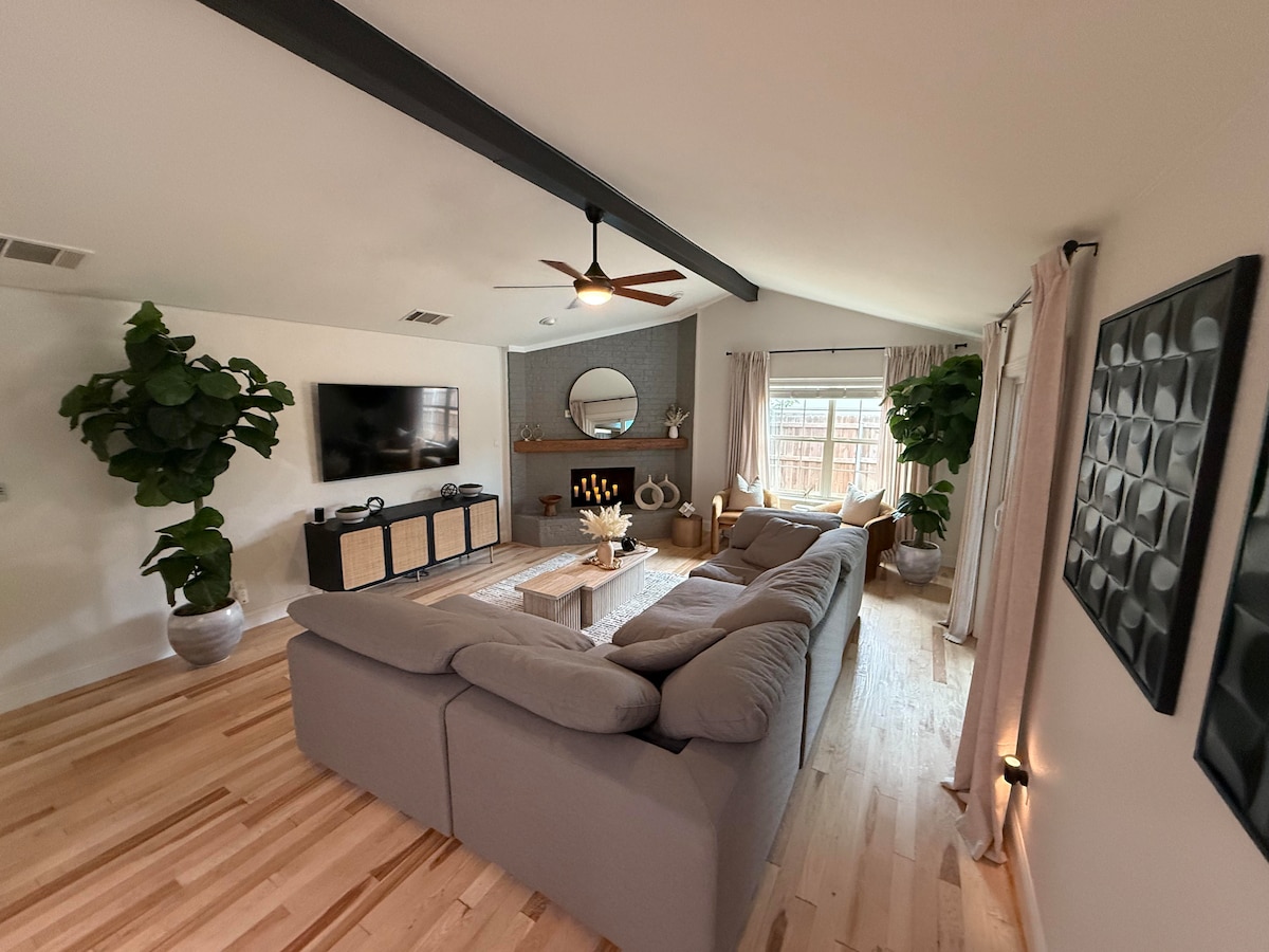 The living area features a spacious sectional sofa arranged around a central coffee table. A large wall-mounted TV is positioned above a stylish console. Natural light filters through the window, complemented by soft drapery. Potted plants add a touch of greenery to the warm, wooden flooring.
