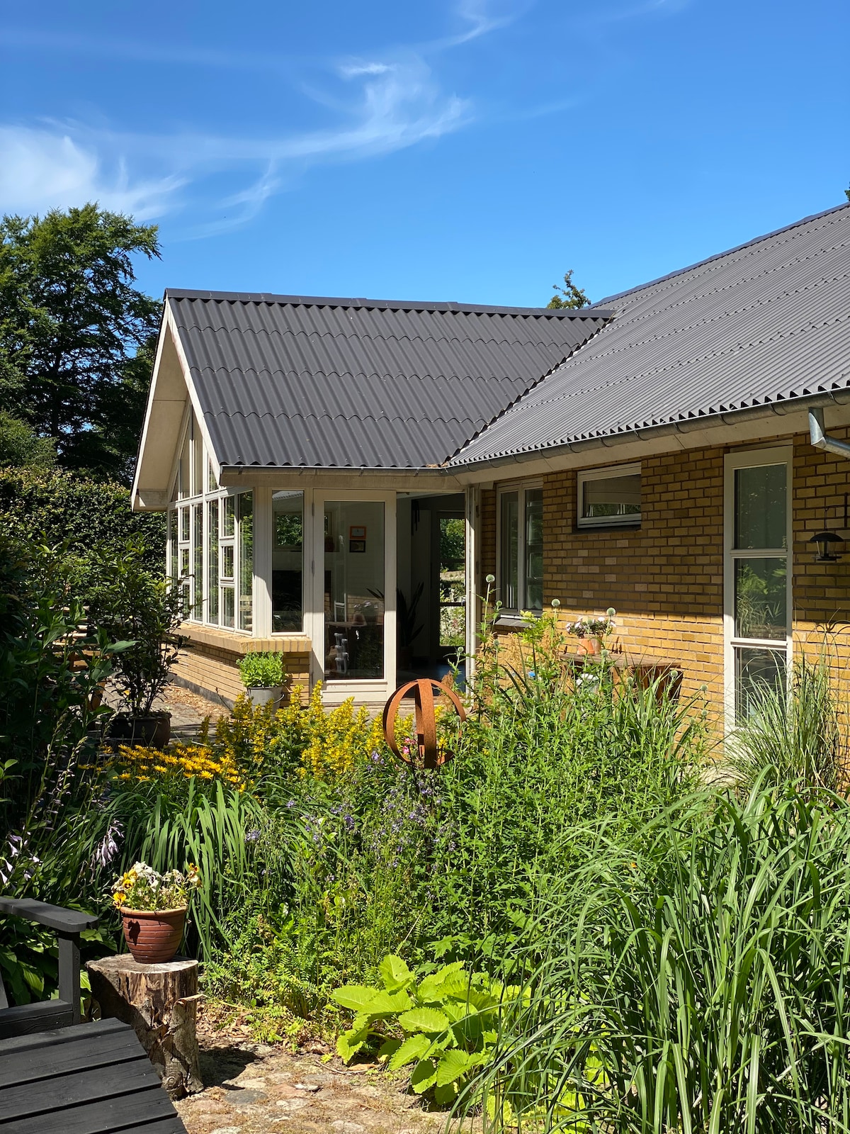 A sunny view of the home’s exterior is presented, showcasing a blend of brick and large glass windows. Lush greenery and colorful flowers surround the cozy patio area, where a decorative pot enhances the garden's natural beauty.