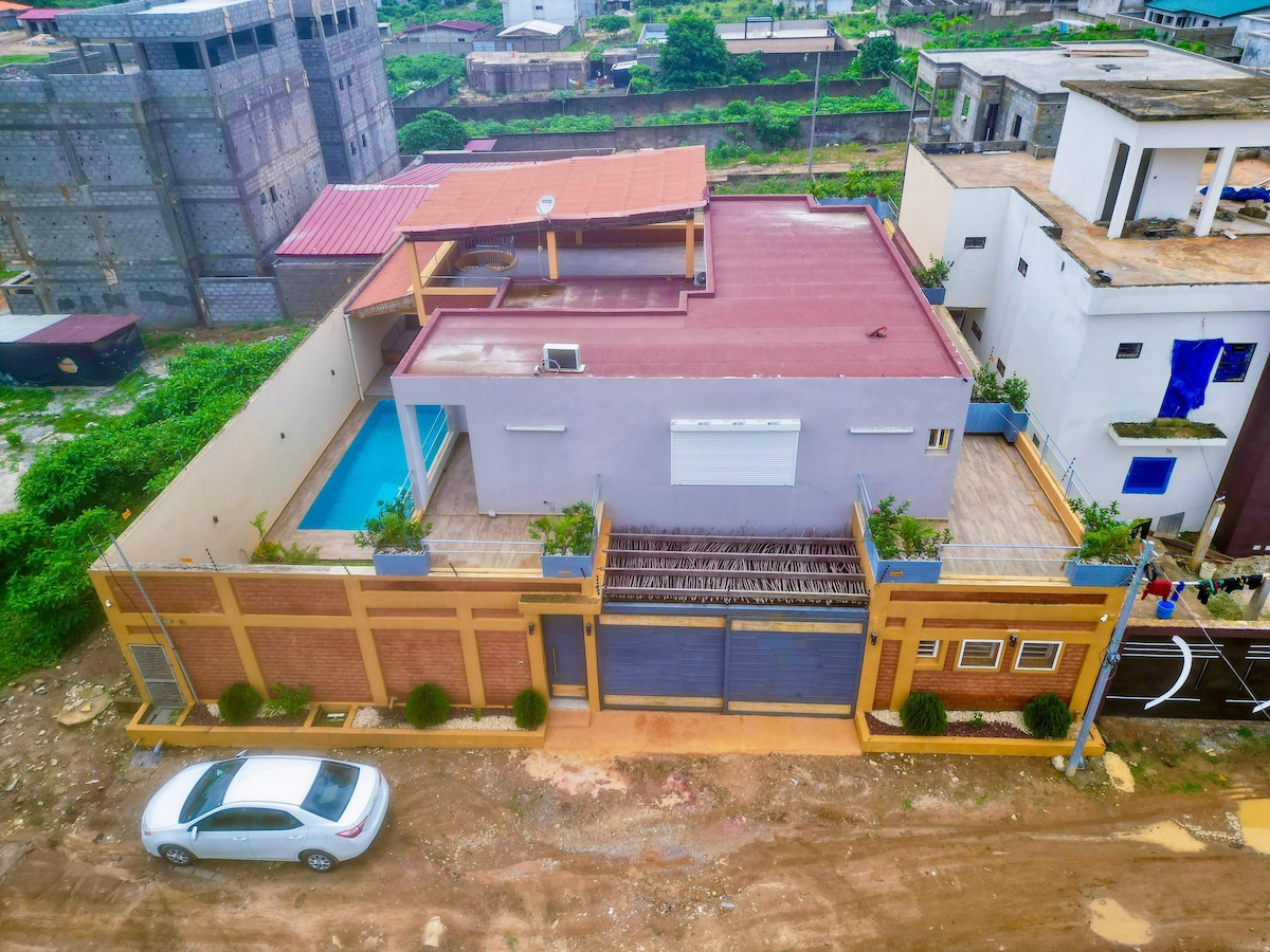An aerial view showcases a modern villa with a red rooftop and surrounding greenery. A private pool is visible in the backyard, with well-maintained plants lining the entrance. The design emphasizes open areas complemented by surrounding residential structures.