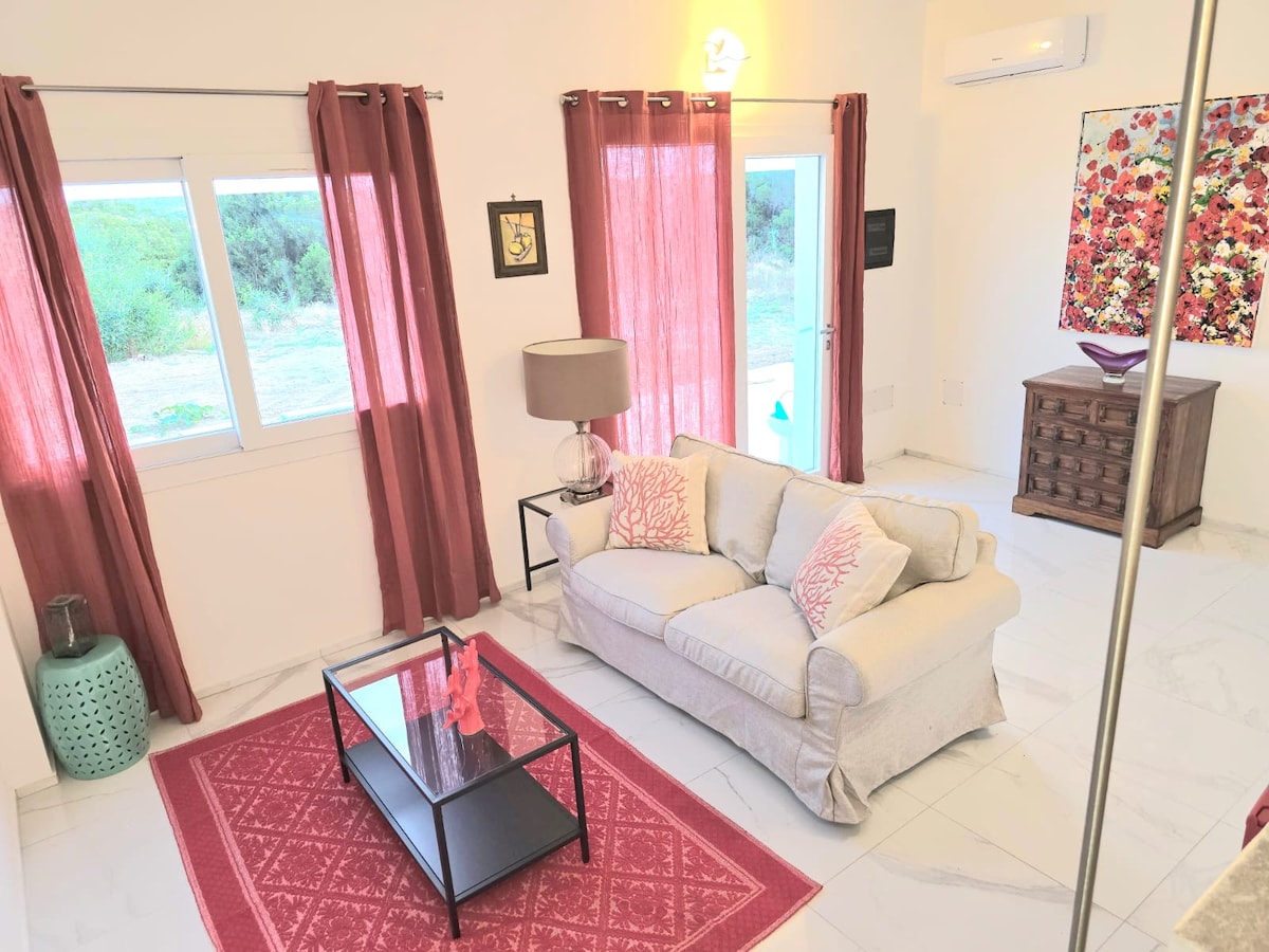 A light-filled living area features a comfortable sofa adorned with decorative pillows, set against a backdrop of vibrant curtains. A coffee table sits in the center, while artwork and a lamp add visual interest. Large windows offer views of the surrounding greenery.