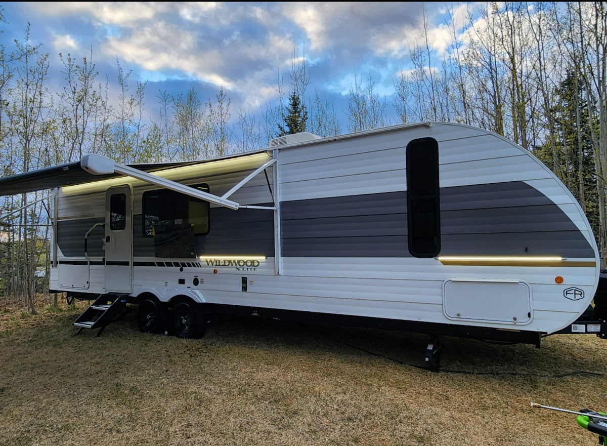 A modern travel trailer is situated on a grassy area, surrounded by trees. Its sleek exterior features a mix of white and gray panels, with an extended awning providing shade. Soft lighting accents the contours of the trailer, enhancing the outdoor space.