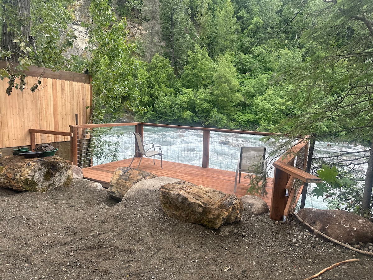 A wooden deck is positioned at the river's edge, featuring two camp chairs and a propane grill. Natural boulders and sandy ground encompass the area, while lush greenery provides a backdrop. The rushing water of the river is visible beyond the railing.
