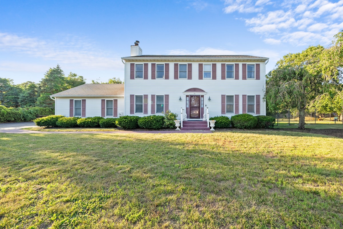 A two-story house is set on a spacious lawn, surrounded by greenery. The facade features multiple windows with red shutters, and a central front door is framed by two white columns. A stone pathway leads to the entrance, with a clear blue sky above.