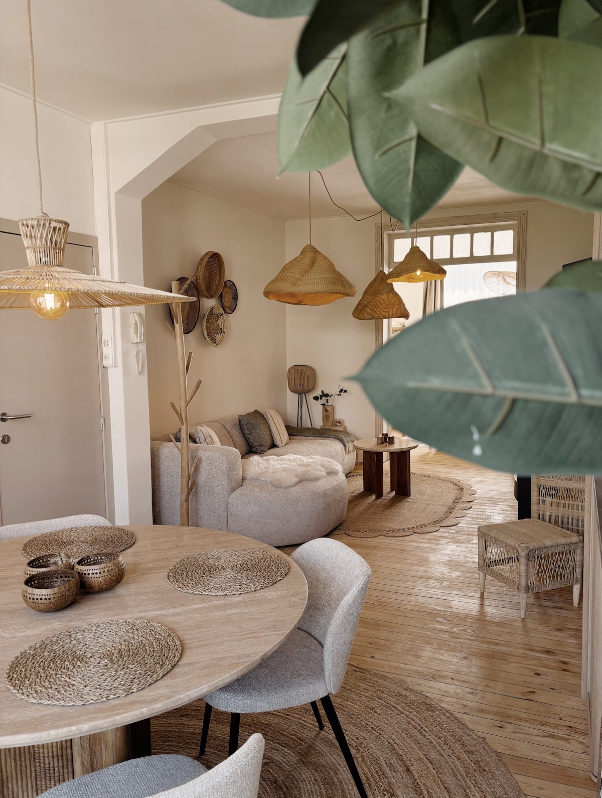 A cozy dining area features a round wooden table surrounded by light-colored chairs. Natural textures are highlighted with woven placemats and decorative bowls. A soft seating area is visible in the background, with textured pillows and pendant lights adding to the inviting ambiance.