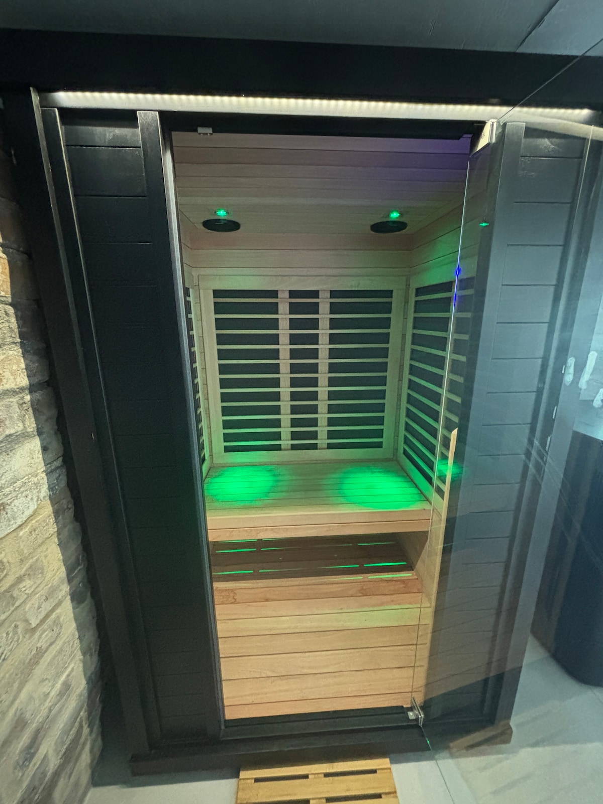 An indoor full-spectrum infrared sauna is displayed, featuring wooden slats and ambient green lighting. The glass door allows a view of the spacious seating area, ensuring comfort during use. The surrounding stone wall further complements the sauna's modern aesthetic.