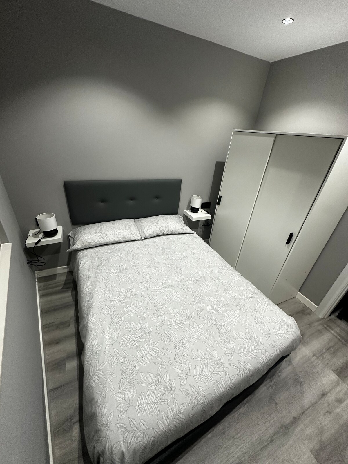 A tranquil bedroom features a spacious bed with light-patterned bedding. Two nightstands with lamps are positioned on either side. A minimalist wardrobe is present, complementing the subtle grey walls and modern flooring, creating an uncluttered and restful environment.