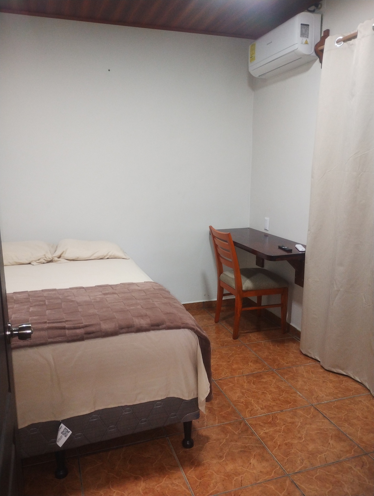 The room features a compact arrangement with a spacious bed dressed in neutral linens. A small wooden desk and chair are positioned near a wall, with a light-colored curtain covering a window. An air conditioning unit is mounted above the desk.