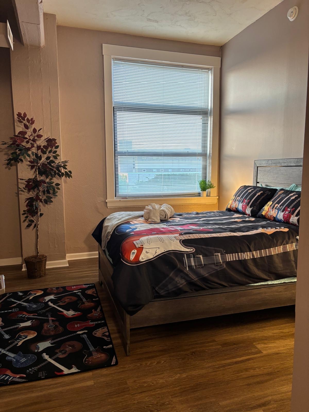 A cozy bedroom features a spacious bed adorned with a guitar-themed duvet cover. Natural light filters through the window, illuminating the room. A small potted plant rests on the windowsill, while a guitar-patterned rug adds a playful touch to the wooden floor.
