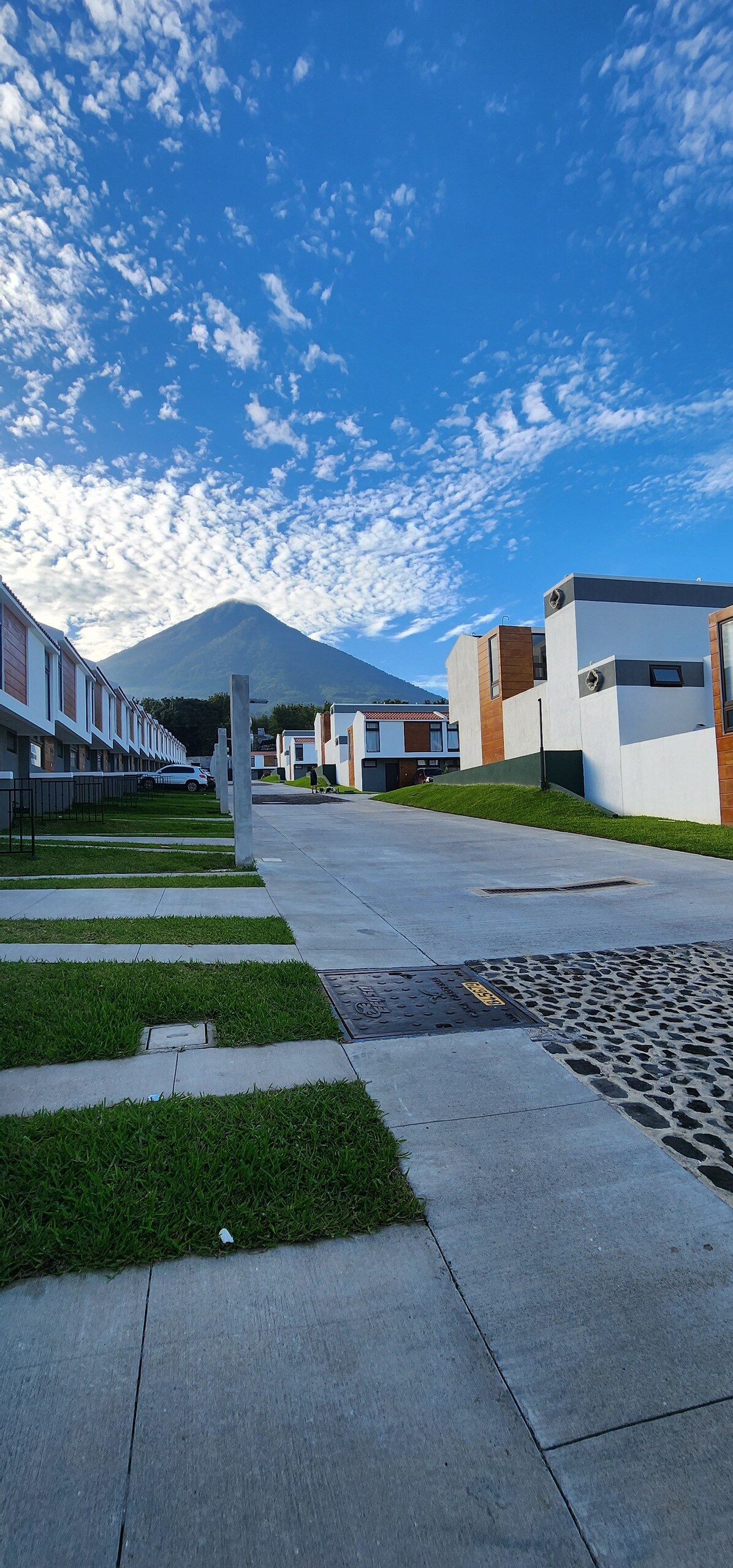 A serene view of the neighborhood showcases modern homes along a clean pathway, framed by well-maintained green lawns. A prominent volcano rises in the background under a clear blue sky, contributing to the peaceful atmosphere.