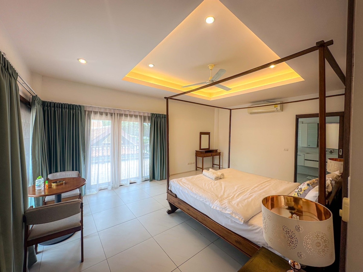 A spacious bedroom is highlighted by a large four-poster bed, white linens neatly arranged. Natural light floods the room through expansive glass doors, flanked by floor-to-ceiling curtains. A small dining table and chairs are positioned nearby, while a cozy seating area adds to the comfort.