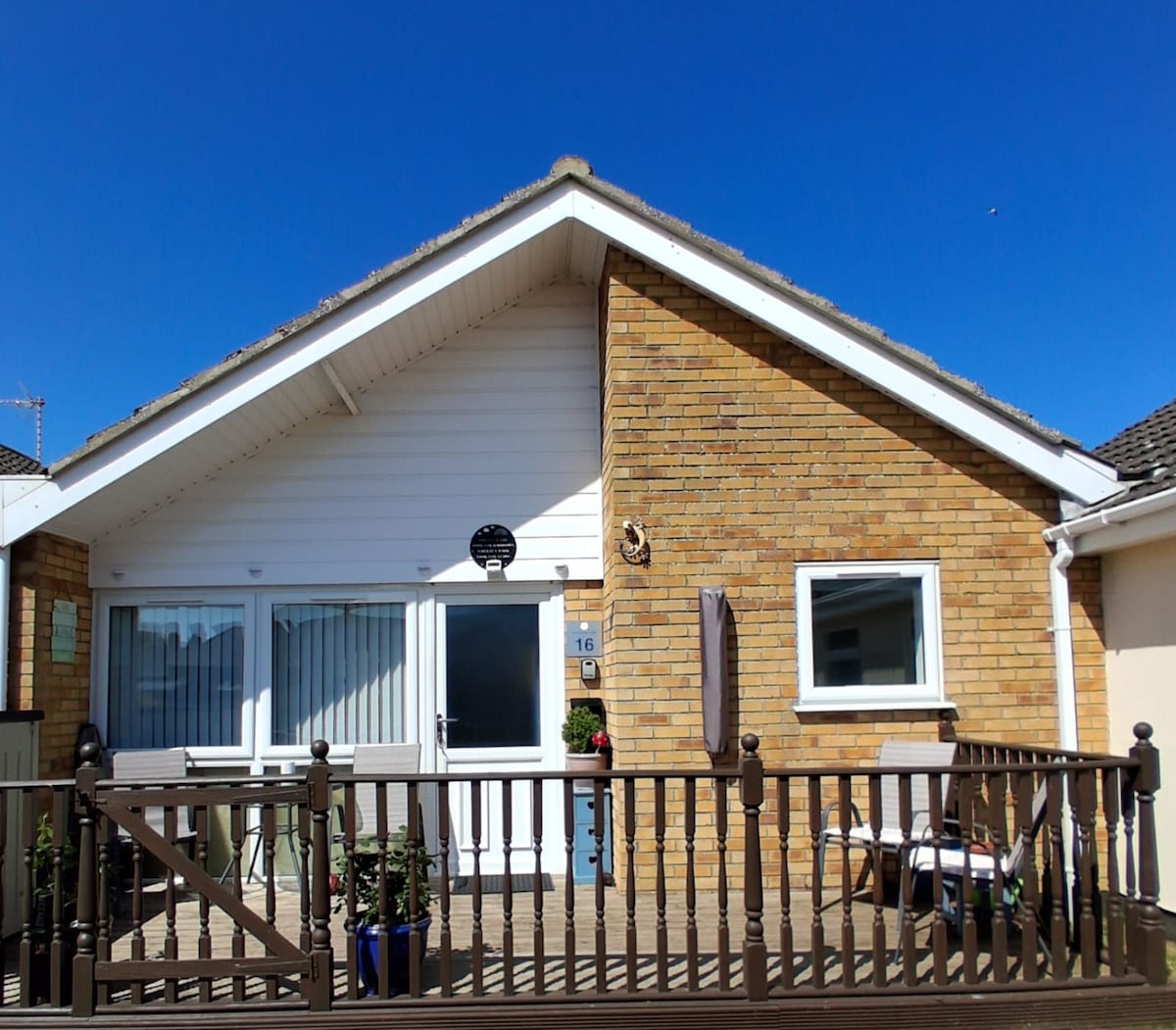 A charming bungalow features a combination of brick and white paneling. Large windows allow natural light to fill the interior. A secure wooden deck is adorned with potted plants, while a small table and chairs await on one side, providing a cozy outdoor space.