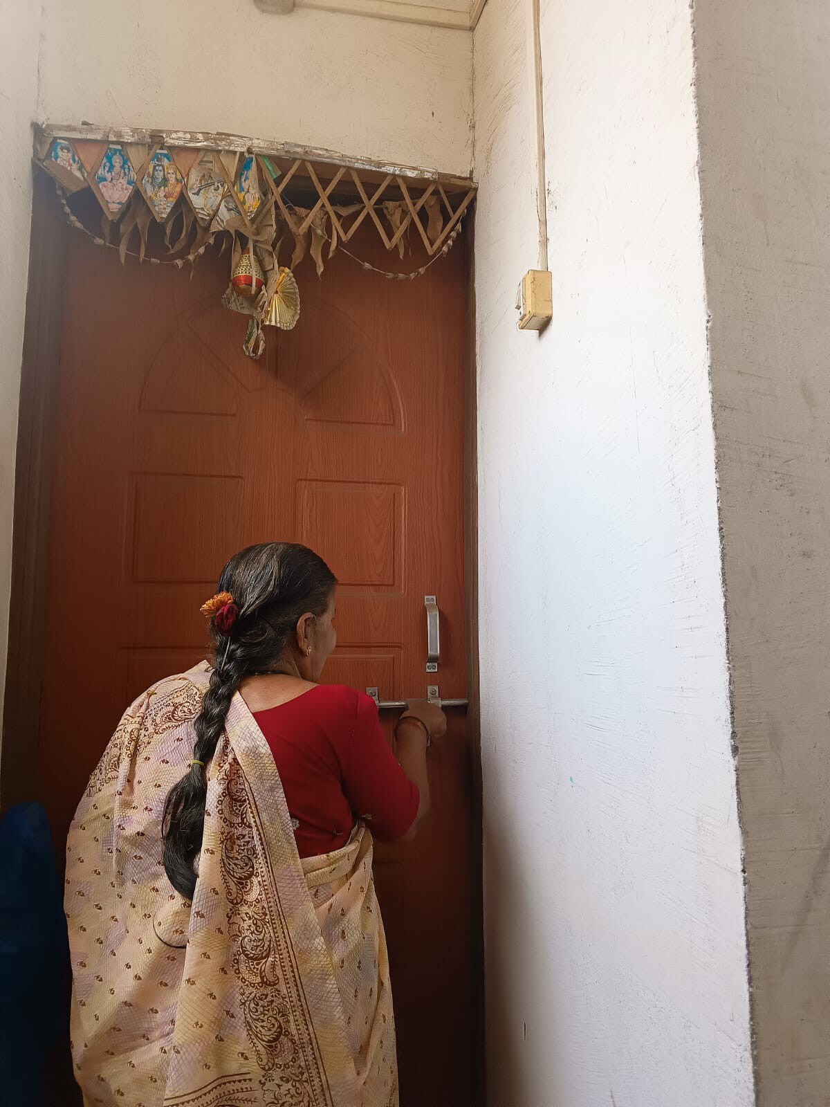 A woman in a traditional saree is shown standing in front of a closed wooden door, locking it. The door features a simple design, and a decorative piece hangs above it, adding a cultural touch to the entrance.