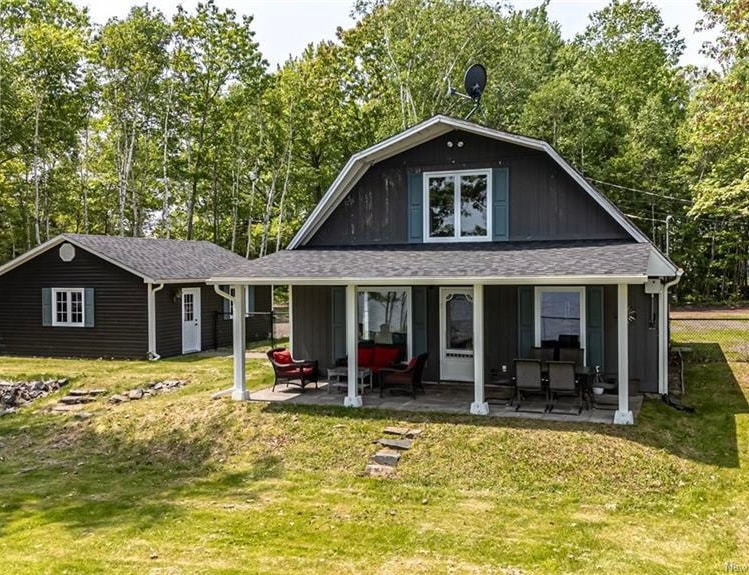 The exterior of the house features a covered porch with seating arrangements. A well-kept yard is visible, bordered by trees and shrubs. Adjacent to the main structure is a separate building, adding to the property’s charm and functionality.