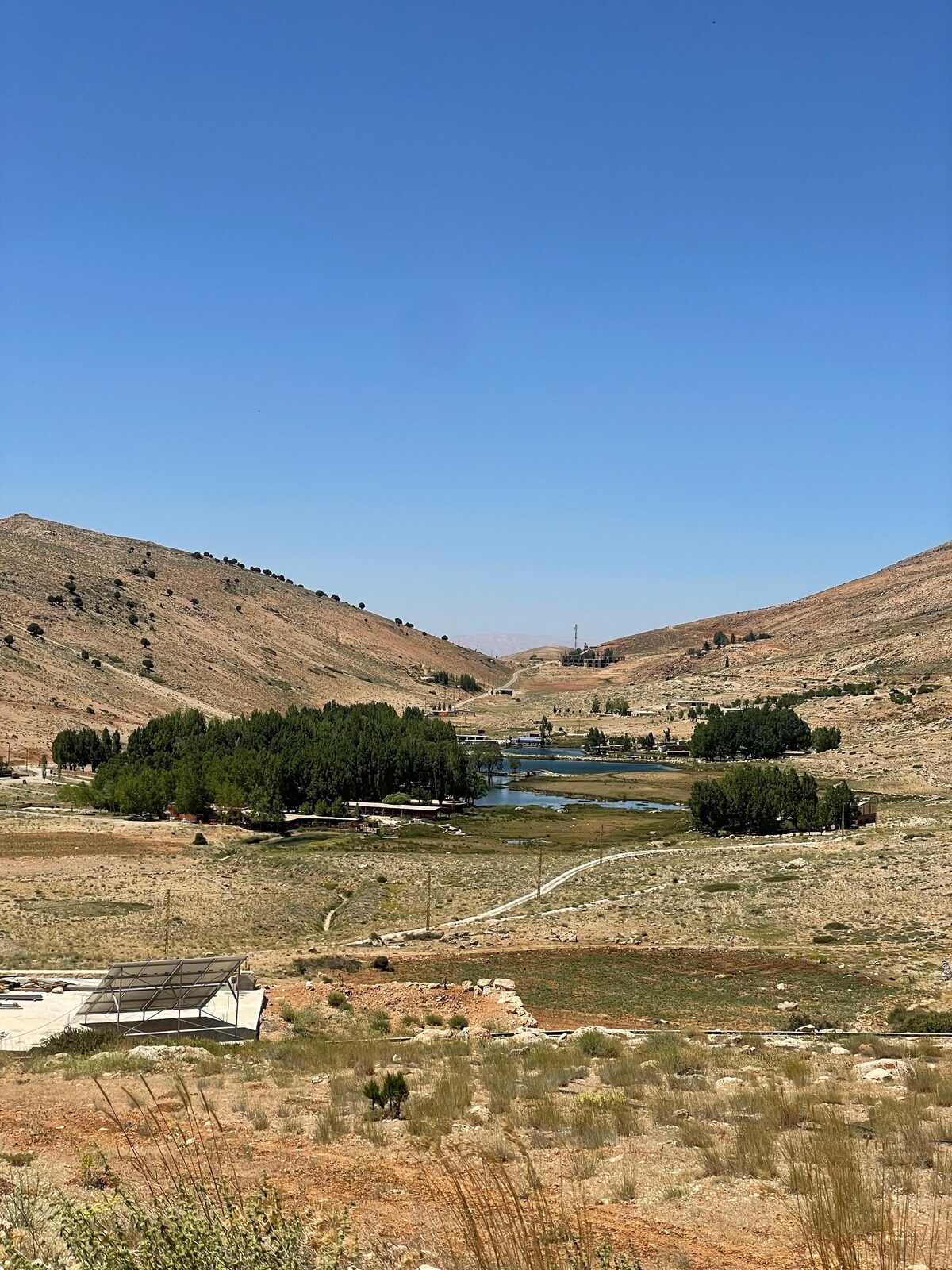 A panoramic view of rolling hills and a tranquil valley is captured under a clear blue sky. Lush green trees line a serene waterway, while a grassy expanse stretches across the landscape. The distant mountains create a natural backdrop, enhancing the peaceful scenery.