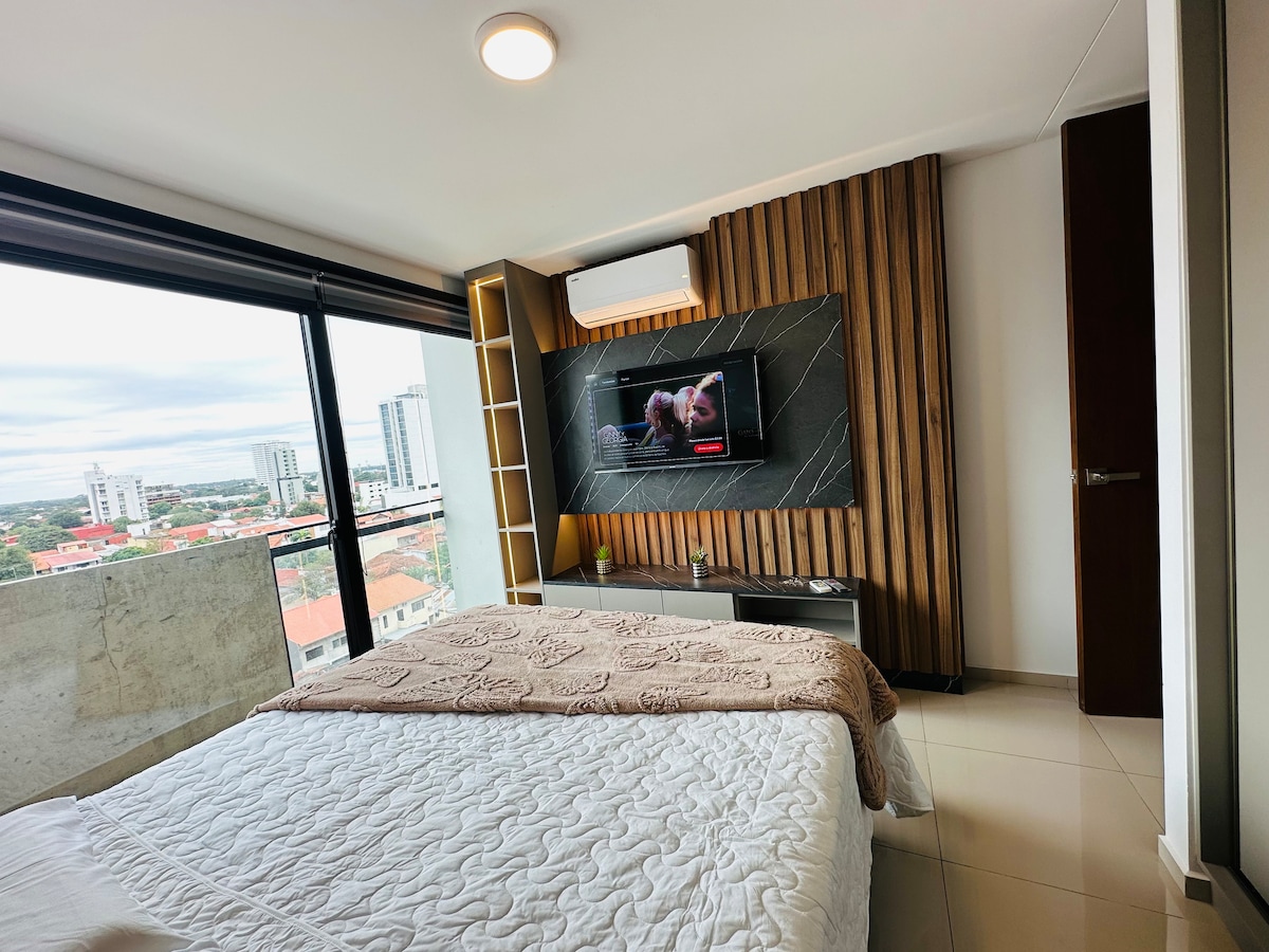 A modern bedroom features a spacious bed covered with a light quilt. A flat-screen TV is mounted on the wall, alongside a decorative wooden panel. Large windows provide city views and ample natural light, while a soft light fixture adds to the ambiance.