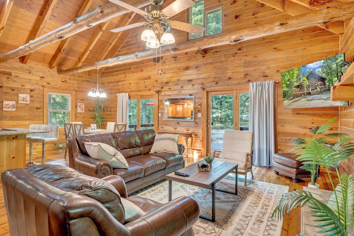 The living area features a cozy arrangement of leather sofas and a coffee table on a patterned rug. Large windows allow for natural light, showcasing the wooden walls and ceiling beams. A flat-screen TV is mounted on the wall, providing a modern touch to the rustic setting.