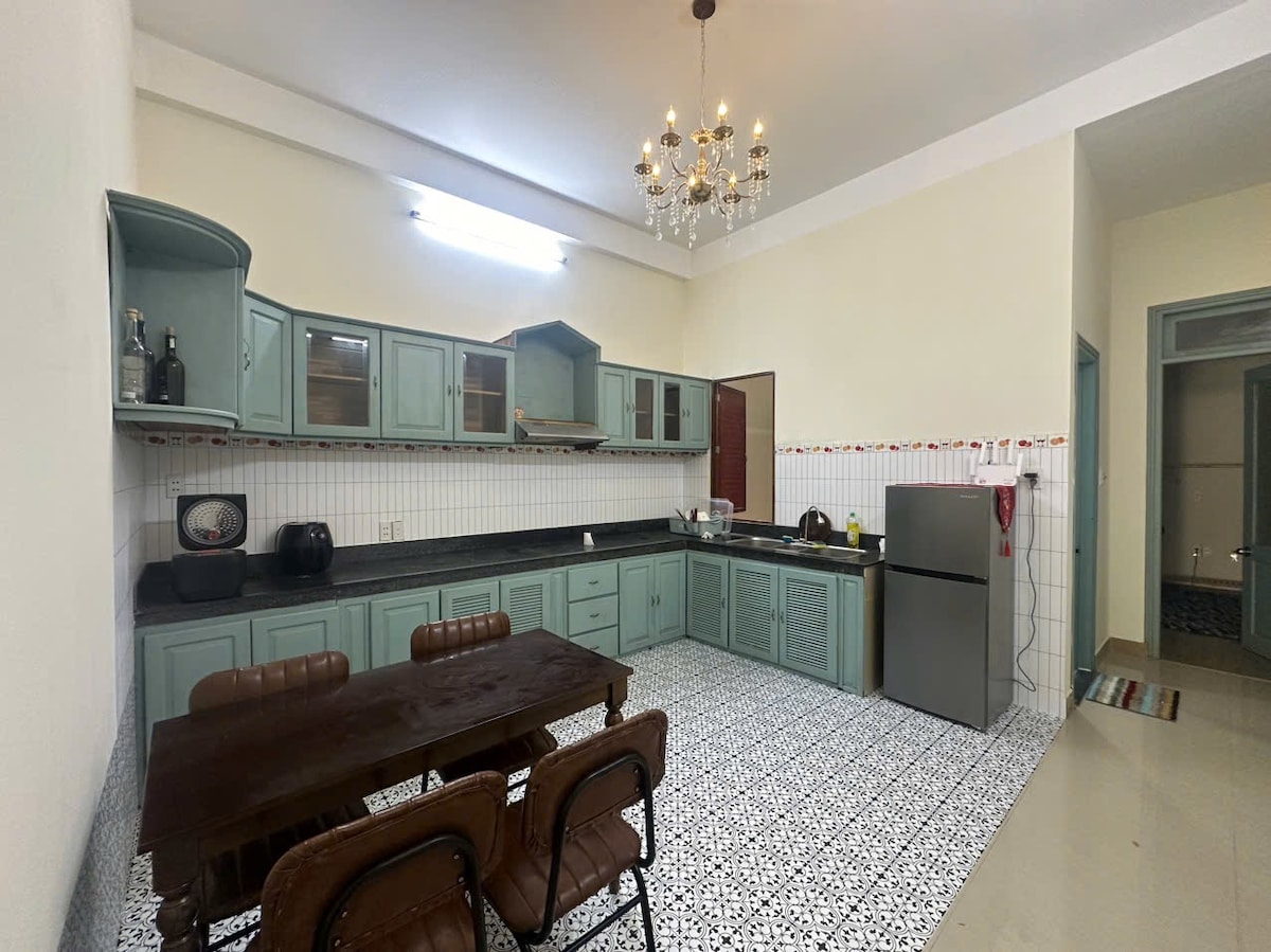 A spacious kitchen features a large dining table surrounded by four chairs. Light blue cabinetry complements the black countertops and a stainless-steel refrigerator. Vintage tiles in black and white cover the floor, while a chandelier provides illumination from above.