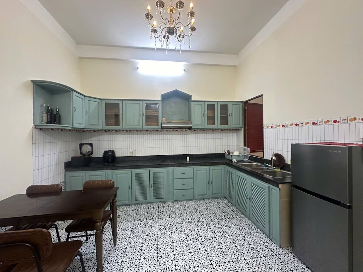 A spacious kitchen is featured, characterized by pale green cabinetry and a dark countertop. A dining table with wooden chairs is visible, while a silver refrigerator stands against the wall. Decorative tiles adorn the floor, providing a distinct pattern.