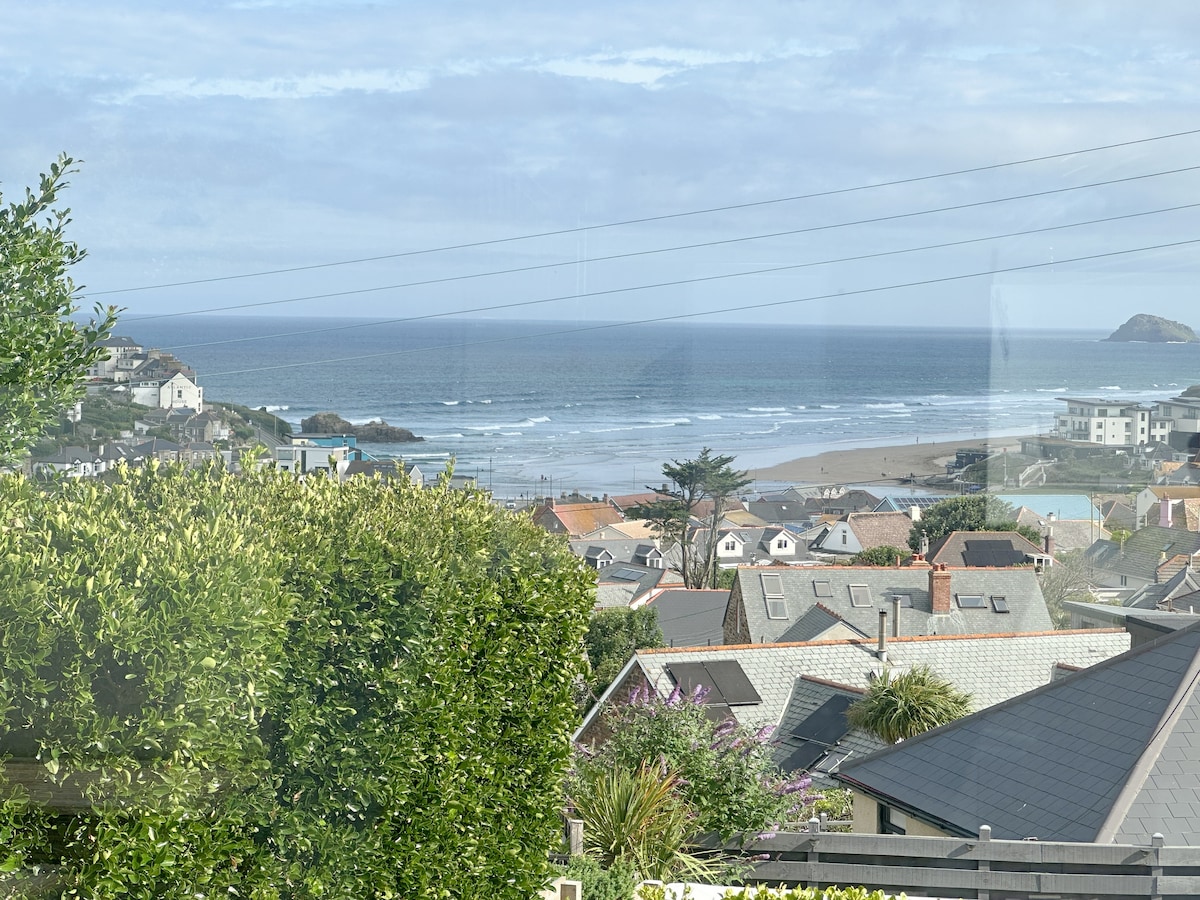 A view showcases the coastline with gentle waves lapping against the sandy beach. Rolling hills and coastal homes dot the landscape, framed by lush greenery. The sky is bright with a few clouds, while the ocean glistens in the distance.