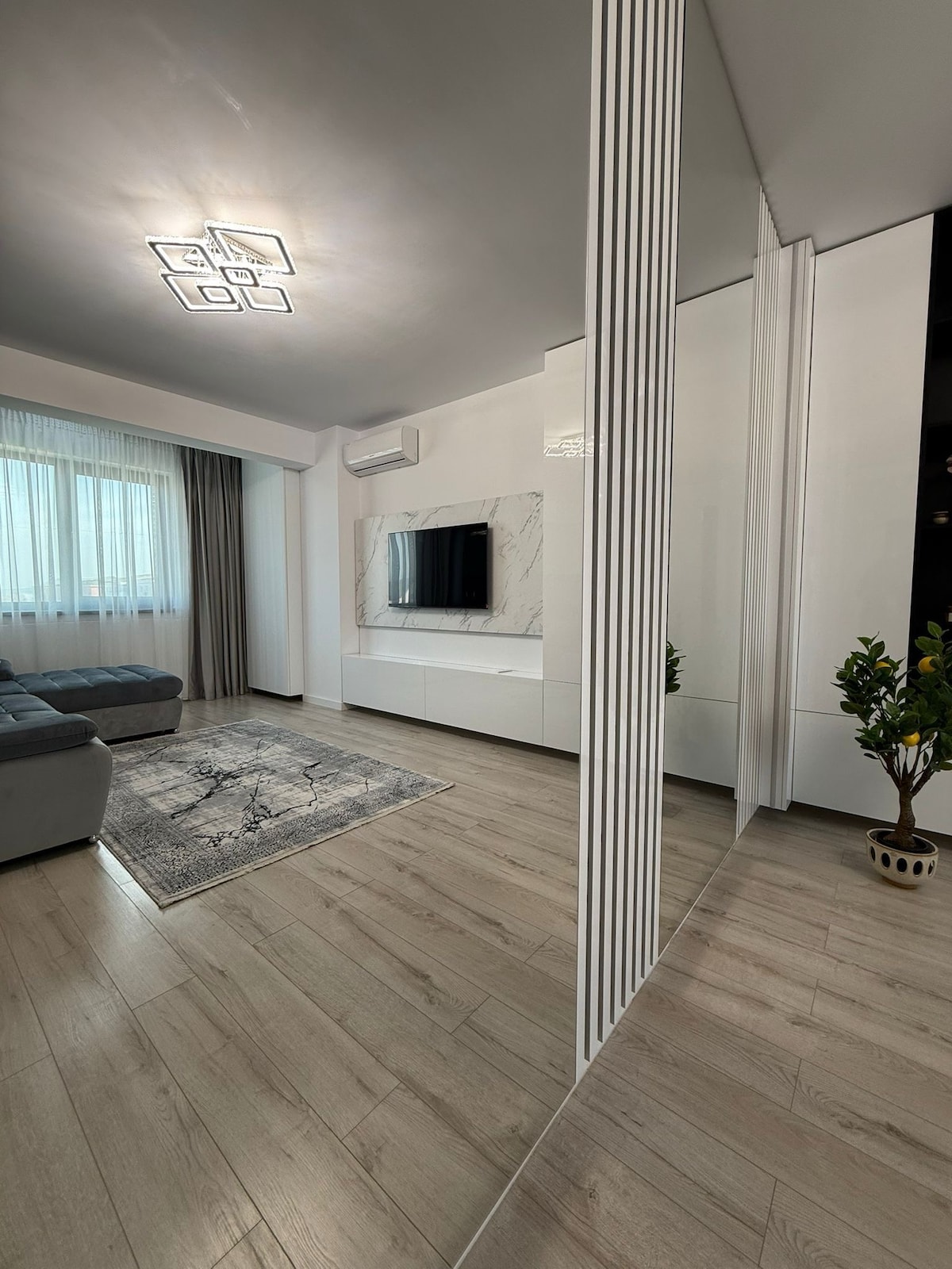 A modern living space is highlighted by light wood flooring and sleek white furniture. A grey sofa is positioned near a decorative rug, and a mounted television is placed on a marble-textured wall. Natural light filters in through the large window adorned with sheer curtains.
