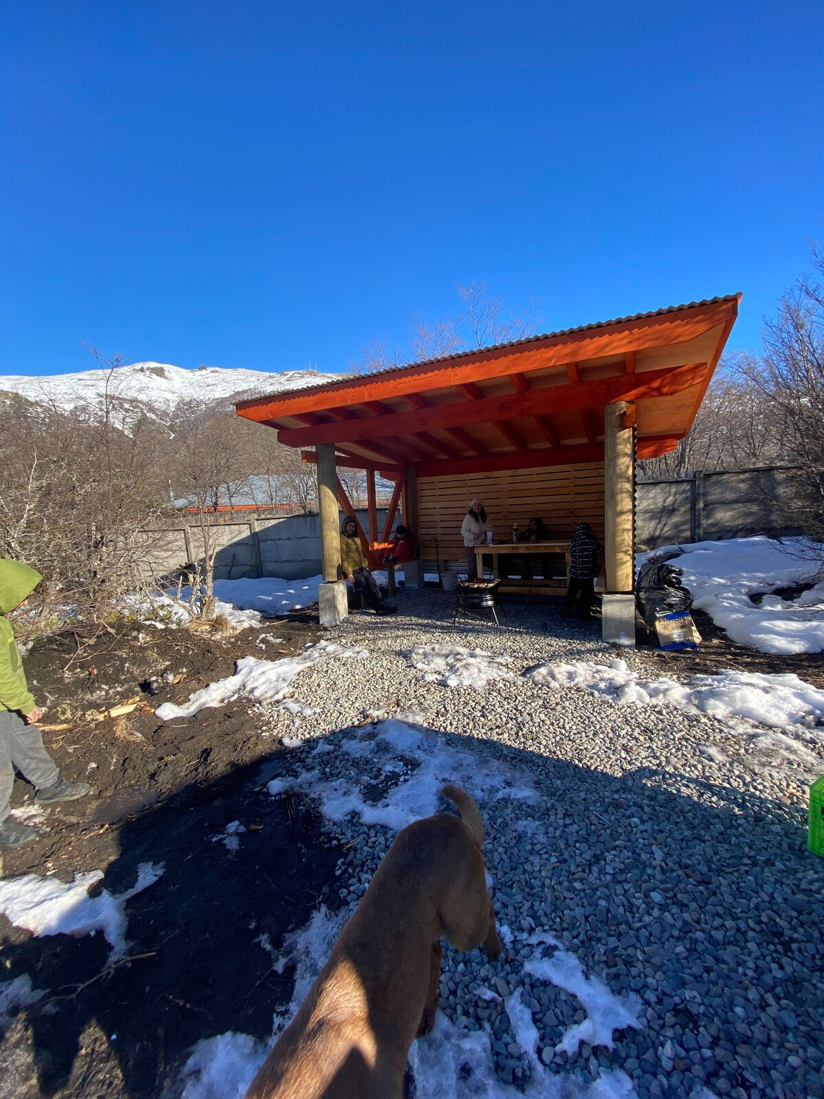 A covered outdoor space is seen, featuring a wooden structure with a vibrant orange roof. A cooking area is set up beneath the shelter, surrounded by gravel and snow on the ground. Sparse vegetation and mountains are visible in the background under a clear blue sky.