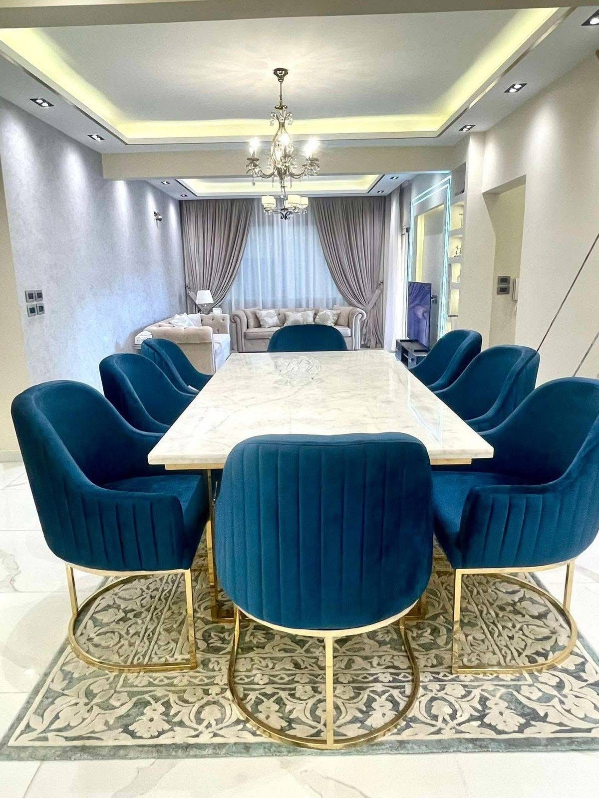 A dining area is highlighted by a long marble table surrounded by eight upholstered blue chairs with golden legs. A patterned rug lies underneath, and a chandelier hangs from the ceiling, illuminating the space. Soft curtains adorn the window, complementing the neutral wall tones.