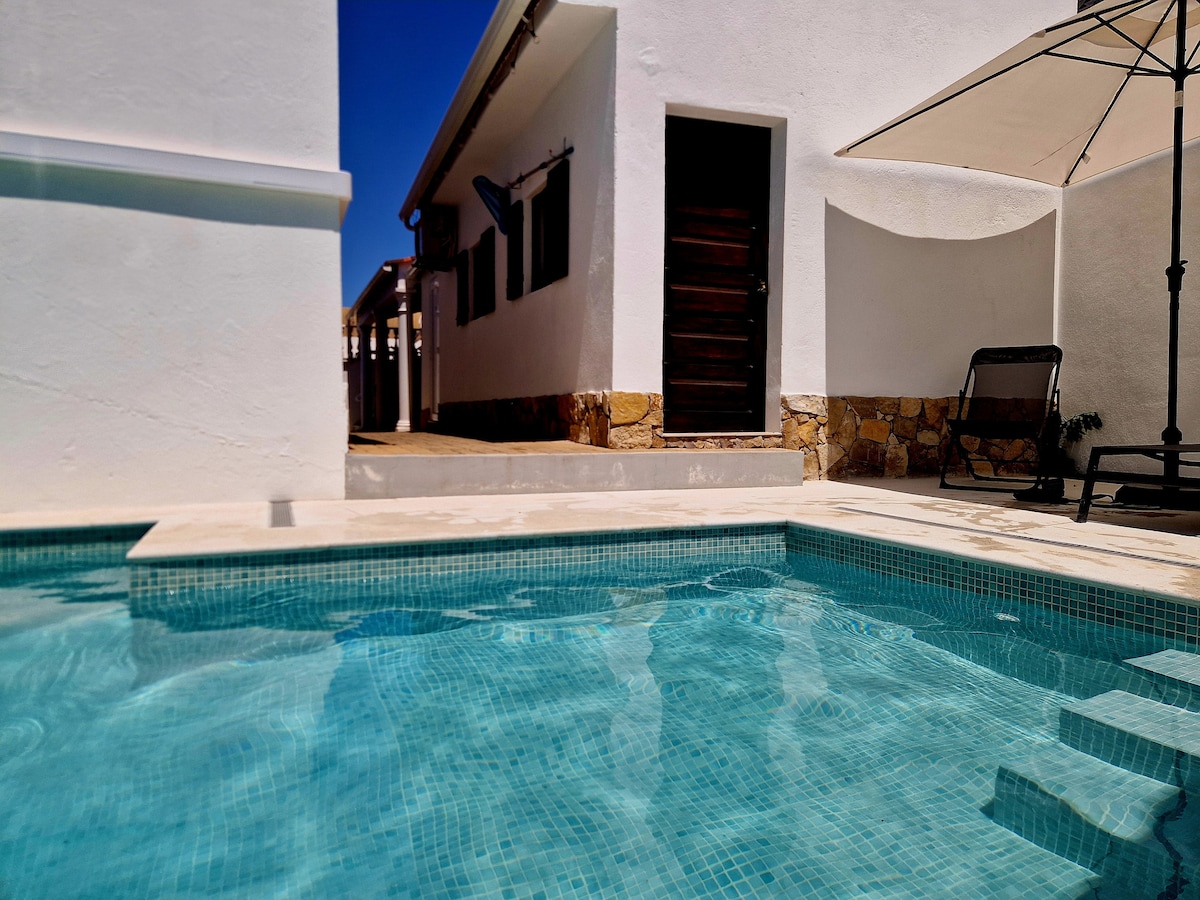 A clear view of a tranquil pool, reflecting light beneath a bright sky. The pool is surrounded by a simple, white facade and stone details. A sun lounger is positioned near the pool, and an umbrella provides shade, enhancing the outdoor space's functionality.