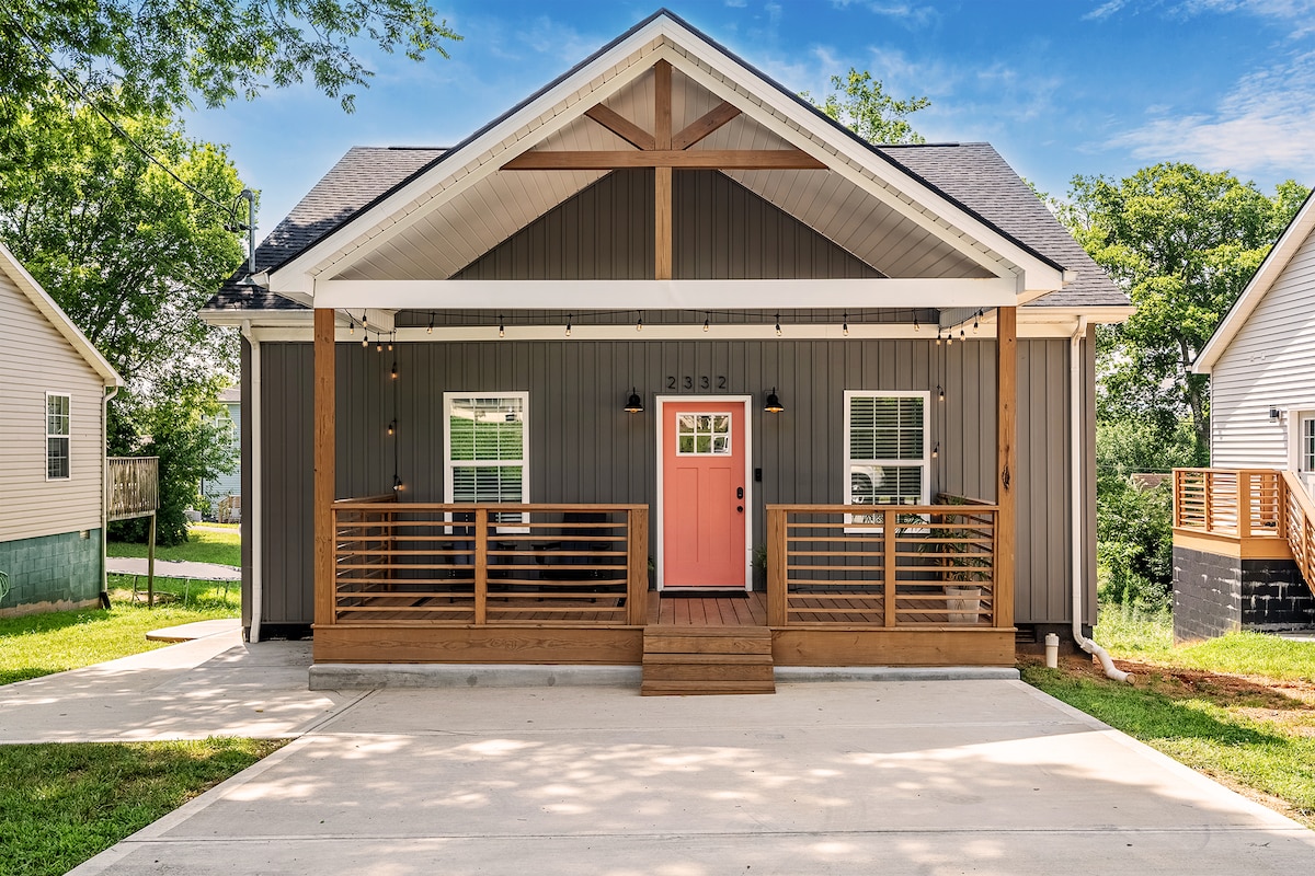 A modern, one-story cottage features a wide front porch with stylish wooden railings and a distinctive pink front door. The house is accented by light grey siding and wooden beams, set against a backdrop of greenery. A spacious driveway leads up to the entrance.