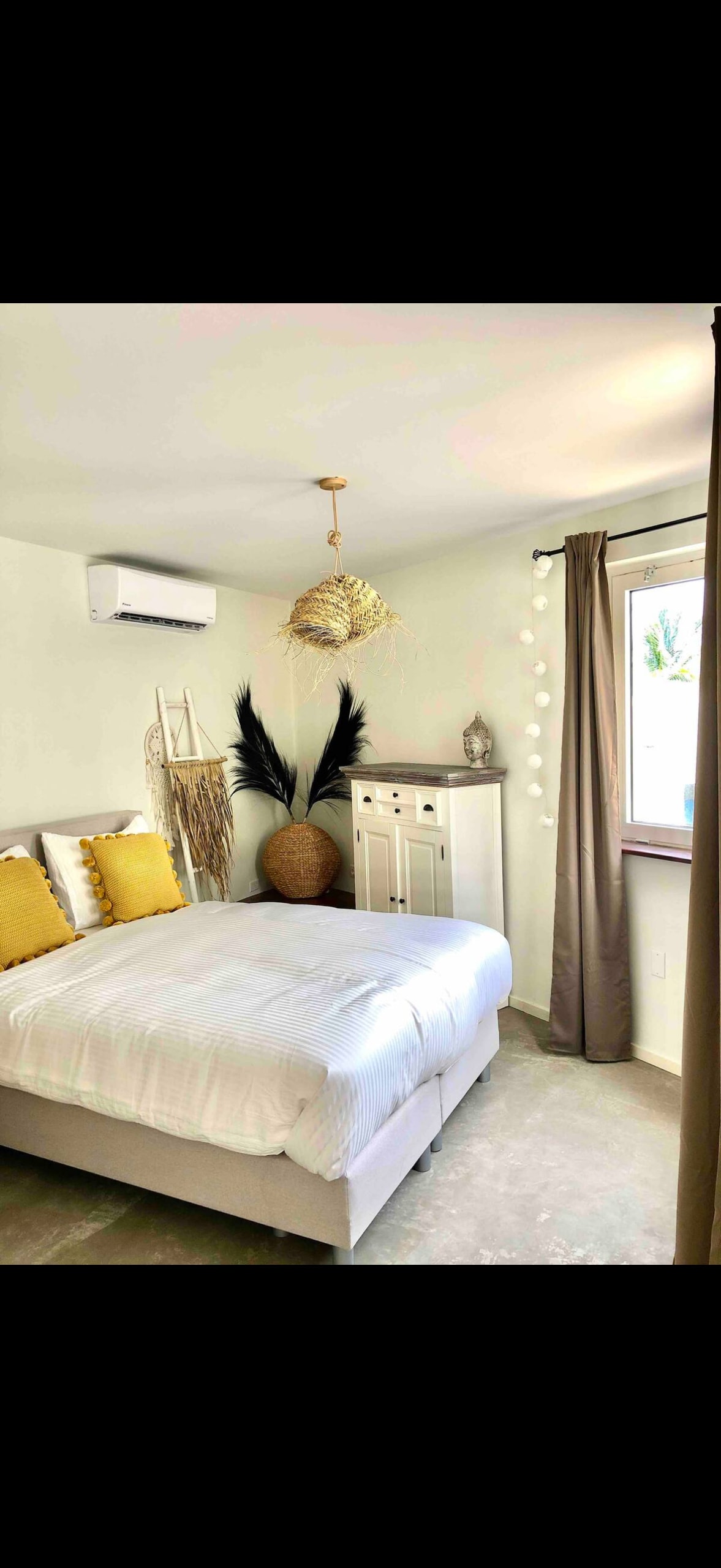A bright sleeping area features a comfortable bed with white linens and yellow accent pillows. A decorative plant arrangement and woven light fixture add texture. A chest of drawers is positioned against the wall, while a window allows natural light to enter, complemented by light curtains.