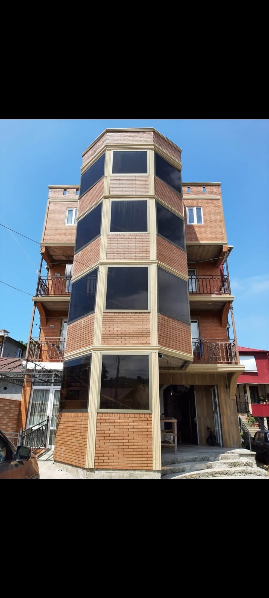 A multi-story brick building is displayed, characterized by large, dark-framed windows that create a modern appearance. The design features a rounded corner, contributing to the structure's unique silhouette against a clear blue sky. Balconies are visible on each floor, enhancing the exterior.