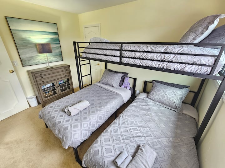 - Bunk room setup with full bed below, two twin beds, and matching linens for a restful night
- Nautical wall art & soft yellow walls add a cheerful coastal vibe to the space
- Compact dresser & soft lighting create a cozy family friendly sleep area